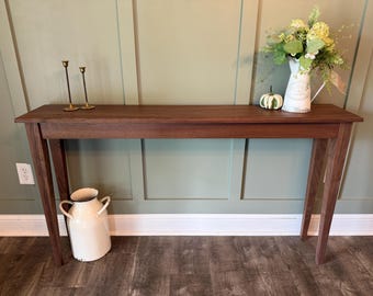 Op de afbeelding: Een lange, rechthoekige houten sidetable met vier taps toelopende poten. Op het tafelblad staan twee dunne koperen kandelaars, een kleine witte pompoen en een witte kan gevuld met groen blad. Een grote witte metalen melkbus staat eronder.