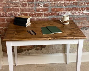 Op de afbeelding: Een witte houten tafel met een bruin gebeitst blad. Op de tafel ligt een stapel notitieboekjes, twee groene notitieboekjes, drie pennen en een keramische mok.