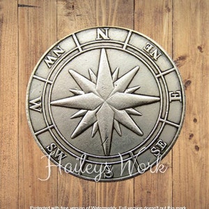 Puede incluir: Una placa de pared circular de metal plateado con forma de rosa de los vientos y una estrella central de ocho puntas. El anillo exterior muestra los puntos cardinales e intercardinales N, NE, E, SE, S, SW, W y NW en relieve.