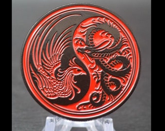 May include: A circular metal coin featuring a black and red yin-yang design. One side displays a detailed black phoenix with intricate wing feathers, while the other side shows a coiled black dragon, both set against a vibrant red background.