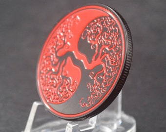 May include: A circular metal coin featuring a detailed red enamel tree design set against a matte black background. The coin has a textured, ridged edge and is displayed at an angle, showcasing the contrast between the vibrant red and black.