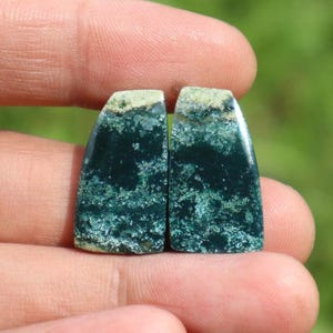 May include: Two polished gemstones with a dark teal base color and a speckled, light green pattern. The top edge of each stone has a band of light green and tan. The stones are held in a person's hand.