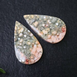 Könnte beinhalten: Zwei tropfenförmige, polierte, natürliche Stein-Cabochons. Die Steine sind eine Mischung aus Rosa, Weiß und Grau mit einem gesprenkelten Muster.