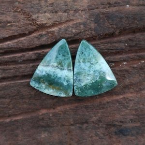 May include: Two polished, triangular gemstones with a mix of green, white, and brown hues. The stones have a smooth, glossy surface and are set against a dark brown, textured wooden background. The gemstones appear to be natural, with unique patterns.