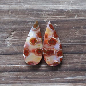 May include: Two teardrop-shaped gemstones with a unique pattern of red, orange, and brown spots on a cream and white background. The gemstones are displayed on a wooden surface, showcasing their natural beauty.