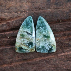 May include: Two polished, teardrop-shaped gemstones with a mix of green, white, and black patterns. The stones are set against a dark brown, textured wooden surface, showcasing their natural beauty and unique color variations.