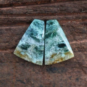May include: Two polished, triangular gemstones with a mix of blue, green, and white hues. The stones have a marbled appearance, with areas of darker green and a touch of yellow at the base. The background is a dark brown, textured surface.