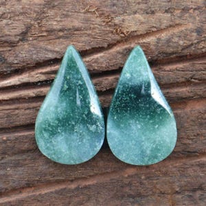 May include: Two teardrop-shaped gemstones with a gradient of green hues. The stones have a polished, glossy surface and a speckled texture. The background is a textured, dark brown wooden surface.