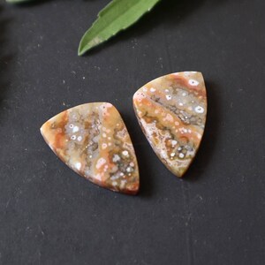 Puede incluir: Dos cabujones de ágata de forma triangular, pulidos, con un patrón de color marrón, blanco y naranja. Los cabujones están sobre una superficie gris oscura.