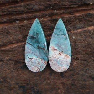 May include: Two teardrop-shaped gemstones with a turquoise and brown marbled pattern. The stones have a polished surface and are set against a dark brown wooden background. These gemstones are likely for jewellery making.