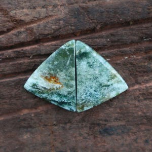 May include: Two matching, triangular-shaped gemstones with a green and white mottled pattern, bisected by a vertical line. The stones have a rough texture and are set against a dark brown, wooden background.
