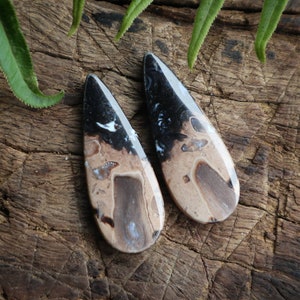 May include: Two teardrop-shaped gemstones with a polished finish. The stones feature a pattern of black, tan, and brown hues. The gemstones are displayed on a rustic wooden surface, with green leaves in the background. These are likely jewelry components.