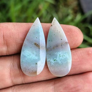 Puede incluir: Dos gemas en forma de lágrima con un acabado pulido. Las piedras muestran una mezcla de tonos azul pálido, verde y marrón, con un patrón en capas. Las gemas se sostienen en una mano, mostrando su belleza natural y variaciones de color únicas.
