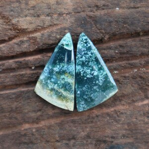 May include: Two triangular-shaped gemstones with a mix of teal, green, and white patterns. The stones have a polished surface and are set against a dark brown wooden background. The gemstones are likely used for jewelry or decorative purposes.