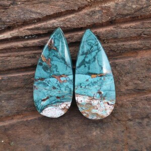 May include: Two teardrop-shaped gemstones with a turquoise and brown pattern. The stones have a polished, glossy finish and a white base. The gemstones are displayed on a brown wooden surface.