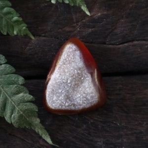 May include: A triangular, red agate geode with a white, sparkly crystal interior. The geode is sitting on a dark brown wooden surface.