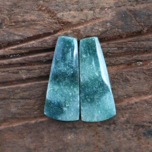 May include: Two polished, trapezoid-shaped gemstones with a gradient of green hues. The stones have a smooth, glossy surface and a semi-translucent appearance. The color transitions from a deep, dark green to a lighter, almost white shade.