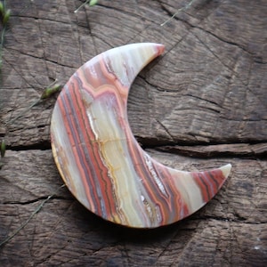 May include: A crescent moon shaped carving made of banded agate with red, white, and brown stripes. The stone is polished and smooth.