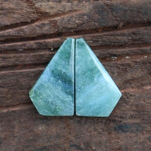 May include: Two polished, triangular-shaped gemstones with a light green and blue color gradient. The stones are displayed on a dark brown, textured wooden surface. The gemstones have a smooth, reflective surface.