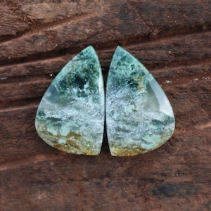 May include: Two teardrop-shaped gemstones with a mix of green, white, and brown hues. The stones have a translucent quality, with a cloudy, layered appearance. The background is a dark brown, textured surface.