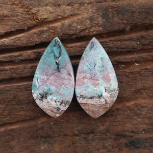 May include: Two teardrop-shaped gemstones with a mix of colors. The stones feature a blend of turquoise, purple, and brown hues, with intricate veining patterns. The bottom edges have a sandy appearance, and the background is a textured wood.