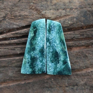May include: Two polished, trapezoid-shaped gemstones with a mottled pattern of teal and dark green. The stones are displayed against a rustic, weathered wooden surface, highlighting their natural beauty and unique textures.
