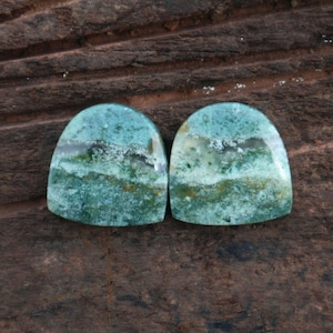 May include: Two polished, cabochon-cut gemstones with a rounded top and a flat bottom. The stones display a mix of teal, green, and white colors, with a layered, natural pattern. The background is a dark, textured wood surface.