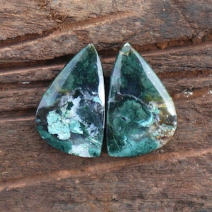 May include: Two teardrop-shaped gemstones with a mix of teal, dark green, and white patterns. The polished stones have a glossy finish and are set against a dark brown wooden surface. The gemstones are likely for jewelry making.
