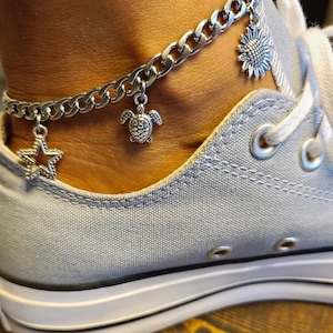 May include: A silver chain ankle bracelet with a star, turtle, and sunflower charm. The bracelet is worn on a person's ankle above a pair of light blue canvas shoes.