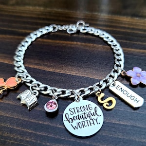 May include: A silver chain bracelet with charms, including a pink heart, a book, a pink crystal, a charm with the text "Strong beautiful worthy", a gold letter "S", and a charm with the text "Enough".