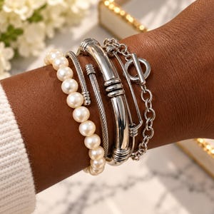 May include: A set of four silver-toned bracelets worn together. The collection includes a strand of white pearls, a twisted metal cable cuff, a smooth polished metal bangle with ribbed accents, and a chain bracelet featuring a circular toggle clasp.