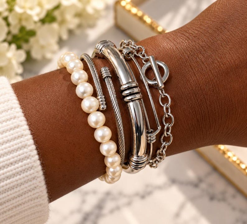 May include: A set of four silver-toned bracelets worn together. The collection includes a strand of white pearls, a twisted metal cable cuff, a smooth polished metal bangle with ribbed accents, and a chain bracelet featuring a circular toggle clasp.