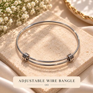 May include: A silver-toned adjustable wire bangle featuring a double-loop design with two sets of three coiled wire accents on each side. The bracelet has a smooth, polished metallic finish and includes the text ADJUSTABLE WIRE BANGLE.