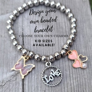 May include: A silver beaded bracelet with charms, including a butterfly, two hearts, and a charm that says "Love". The bracelet is on a wooden surface. The text "Design your own beaded bracelet" and "Choose your own charms" is on the image. The text "Kid sizes available!" is also on the image.