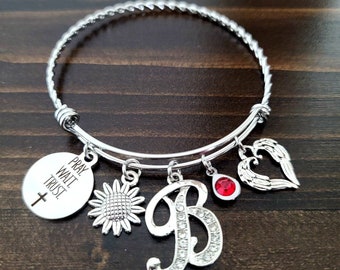 May include: Silver charm bracelet with a variety of charms, including a sunflower, a heart with wings, a red rhinestone, a letter B with rhinestones, and a round charm with the words "Pray, Wait, Trust" and a cross.