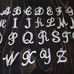 May include: A set of 26 silver charms, each featuring a letter of the alphabet in a cursive script. The charms are adorned with clear rhinestones.