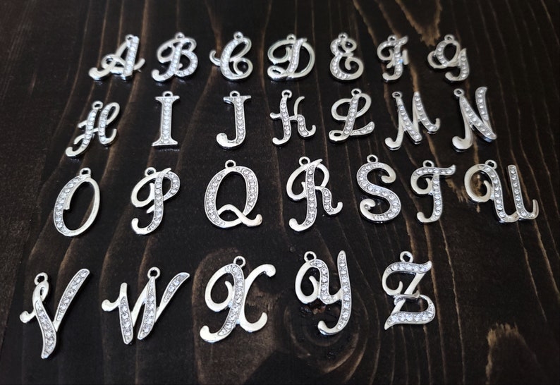 May include: A set of 26 silver charms, each featuring a letter of the alphabet in a cursive script. The charms are adorned with clear rhinestones.
