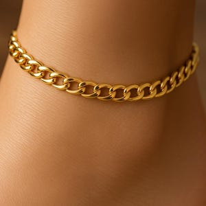 May include: A gold-colored chain anklet. The anklet features a curb chain design, with each link appearing rounded and interlocked. The jewelry is worn around the ankle, and the skin tone is light brown.