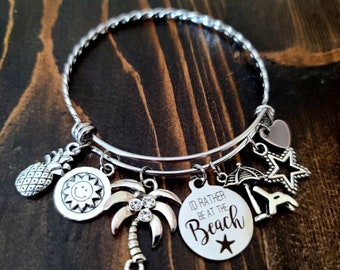 May include: Silver charm bracelet with a variety of beach-themed charms, including a pineapple, a sun, a palm tree, an umbrella, a star, a heart, and a charm that says "I'd rather be at the beach".