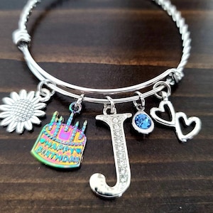 May include: Silver charm bracelet with a rainbow iridescent cake charm that says "Happy Birthday", a silver sunflower charm, a silver letter J charm with rhinestones, a blue rhinestone charm, and two silver heart charms.