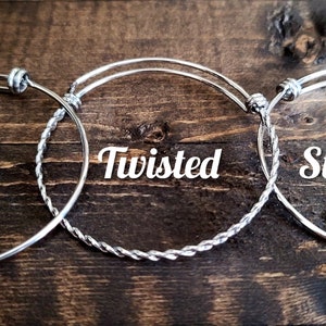 May include: Three silver expandable bracelets with different styles: a standard bracelet, a twisted bracelet, and a stand ball bracelet. The bracelets are on a wooden background.
