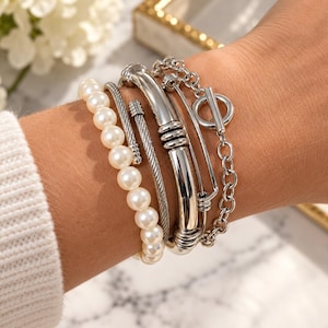May include: A set of four silver-toned bracelets featuring a strand of white faux pearls, a twisted cable wire band, a smooth polished metal cuff with ribbed accents, and a chain link bracelet with a circular toggle clasp closure.