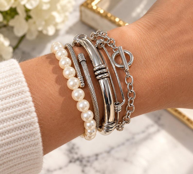 May include: A set of four silver-toned bracelets featuring a strand of white faux pearls, a twisted cable wire band, a smooth polished metal cuff with ribbed accents, and a chain link bracelet with a circular toggle clasp closure.