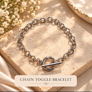 May include: A silver-toned chain toggle bracelet featuring a series of circular links connected to a circular ring and a T-bar closure. The text "CHAIN TOGGLE BRACELET" is printed in black on a light-colored rectangular label below the jewelry.