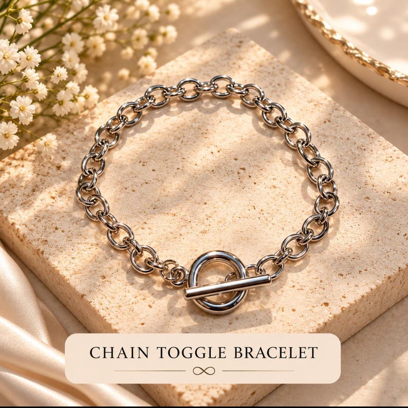May include: A silver-toned chain toggle bracelet featuring a series of circular links connected to a circular ring and a T-bar closure. The text "CHAIN TOGGLE BRACELET" is printed in black on a light-colored rectangular label below the jewelry.