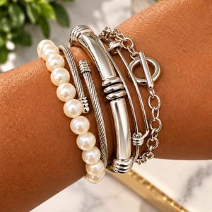 May include: A multi-strand bracelet set featuring a single row of white pearls, a silver-toned twisted cable cuff, a smooth silver-toned metal bar cuff with ribbed accents, and a silver-toned chain bracelet with a toggle clasp closure.
