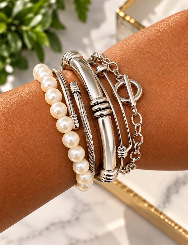 May include: A multi-strand bracelet set featuring a single row of white pearls, a silver-toned twisted cable cuff, a smooth silver-toned metal bar cuff with ribbed accents, and a silver-toned chain bracelet with a toggle clasp closure.
