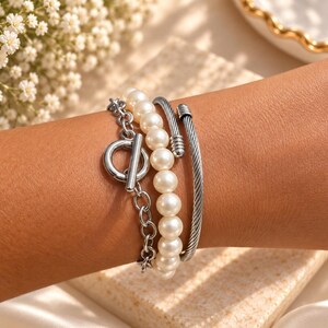 May include: A multi-strand bracelet set featuring a silver-toned chain with a toggle clasp, a strand of white round pearls, and a silver-toned twisted cable cuff. The three distinct bracelets are worn together to create a layered appearance.