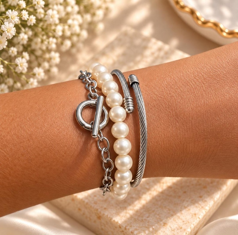May include: A multi-strand bracelet set featuring a silver-toned chain with a toggle clasp, a strand of white round pearls, and a silver-toned twisted cable cuff. The three distinct bracelets are worn together to create a layered appearance.