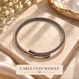 May include: A silver-toned cable cuff bangle featuring a twisted metal wire design. The bracelet has two smooth, cylindrical end caps that overlap slightly. The text CABLE CUFF BANGLE is printed in black on a rectangular label at the bottom.